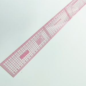 May include: A pink plastic ruler with a grid pattern and inch markings. The ruler is designed for sewing and quilting.