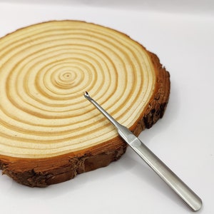 May include: A silver crochet hook with a pointed tip and a wooden base. The hook is lying on a wooden circle with concentric rings.