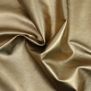 Peut inclure: Gros plan sur un tissu lisse de couleur or, à la texture douce et luxueuse. Le tissu est drapé et plié, créant une impression de mouvement et de fluidité.