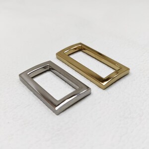 May include: Two metal rectangular rings, one gold and one silver, with rounded edges. The rings are for attaching to a strap or belt.