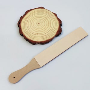 Puede incluir: Una correa de cuero marrón claro con un mango de madera, utilizada para afilar cuchillas. La correa está sobre una superficie de madera.