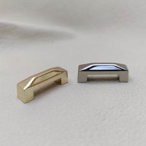 May include: Two small metal drawer pulls, one gold and one silver. Both have a rectangular shape with a raised centre section.