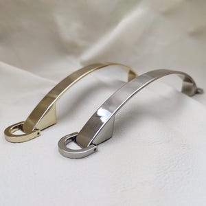 以下が含まれることがあります： 光沢のあるゴールド仕上げとシルバー仕上げの、アーチ型の金属製キャビネットハンドル2点。各ハンドルは湾曲したバーデザインが特徴で、片端にU字型の取り付けベースを備えており、洗練されたモダンな外観を提供します。