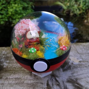 Puede incluir: Una esfera decorativa roja y negra con una parte superior transparente, que presenta un paisaje en miniatura con un personaje rojo y naranja, flores rosas y un arroyo azul. La esfera tiene un botón blanco y se apoya en un soporte transparente.