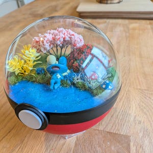 May include: A clear glass terrarium with a red and black base, showcasing a miniature landscape. Inside, a blue figure rests near a blue pond, surrounded by miniature trees, flowers, and a mushroom. The terrarium is set on a wooden surface.