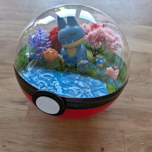 May include: A Snorlax figurine sits inside a clear, spherical display case designed like a red and white Poké Ball. The diorama features a blue water surface, green grass, a pink cherry blossom tree, a purple plant, and small mushrooms.