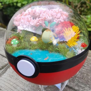 Puede incluir: Un terrario en miniatura dentro de una esfera roja, negra y blanca. La cúpula transparente revela un paisaje en miniatura con un estanque azul, hierba verde, flores de cerezo rosas y un personaje de dibujos animados. Setas amarillas y verdes y plantas coloridas añaden detalles.