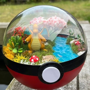 May include: A terrarium inside a red, black, and white Poké Ball. The terrarium features a miniature landscape with a yellow dragon figure, a blue stream, pink flowering trees, and a red mushroom. The Poké Ball sits on a clear stand.