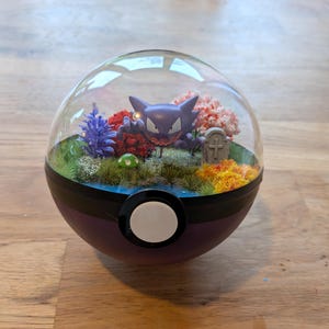 Könnte beinhalten: Eine kugelförmige Vitrine, die einem violett-schwarzen Pokéball ähnelt und ein Miniatur-Diorama enthält. Im Inneren steht eine violette Gengar-Figur auf einem grasbewachsenen Untergrund mit einem kleinen grauen Grabstein, einem grünen Pilz und buntem künstlichem Laub.