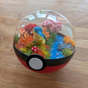 May include: A spherical terrarium featuring a Cyndaquil figure inside a red and black ball. The scene includes a blue stream, a small red and white mushroom, green grass, orange foliage, and a pink cherry blossom tree under a clear dome.