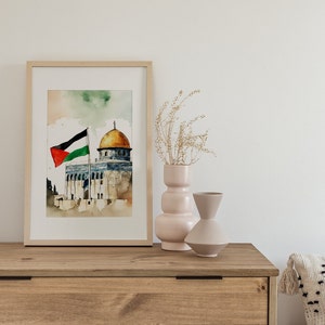 May include: A watercolor painting of the Dome of the Rock in Jerusalem, with a Palestinian flag flying in the foreground. The painting is framed and displayed on a wooden shelf with two decorative vases.