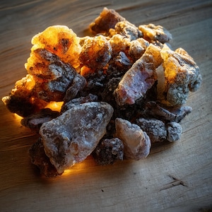May include: A close-up of a pile of amber-colored and gray-toned resin pieces. The resin has a crystalline appearance and is illuminated with warm light, creating a glowing effect. The pieces are resting on a wooden surface.