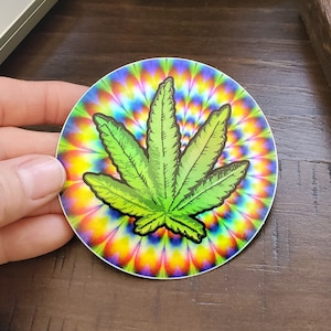May include: A round sticker with a green marijuana leaf on a colorful tie-dye background.