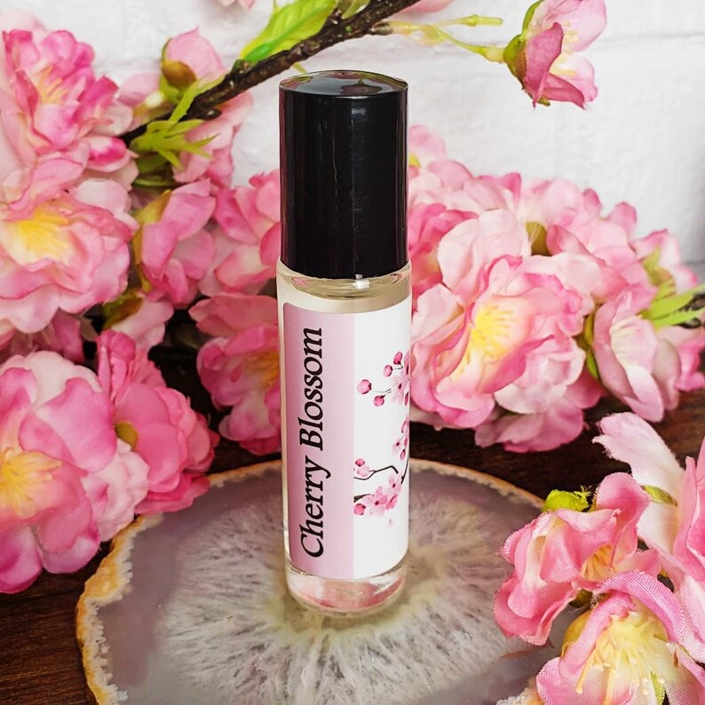 May include: A clear glass roller bottle with a black cap and a white label that says "Cherry Blossom". The bottle is sitting on a white and gray agate slice. The bottle is surrounded by pink cherry blossom flowers.