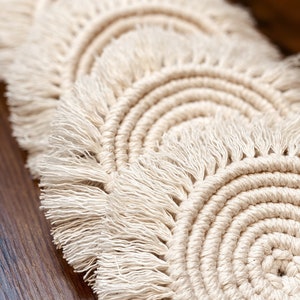 Puede incluir: Conjunto de cuatro posavasos de ganchillo de color crema con detalles de flecos. Los posavasos están dispuestos en forma de semicírculo.