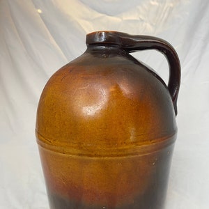 May include: A brown glazed stoneware jug with a wide mouth and a single handle. The jug has a dark brown base and a lighter brown top.