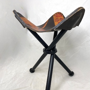 May include: A black metal folding stool with a brown leather seat. The seat has a colorful floral pattern.