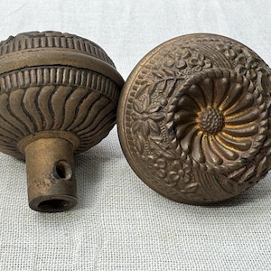 May include: Two antique brass door knobs with intricate designs. One knob has a floral pattern, while the other has a ribbed design.