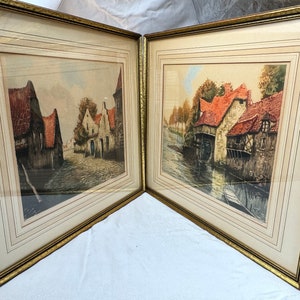 May include: Two framed prints depicting European village scenes. The artwork features buildings with red tile roofs, cobblestone streets, and a canal. The frames are gold-colored with a textured finish, and the prints are behind glass.
