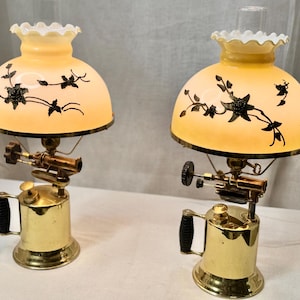 May include: Two antique brass lamps with a unique design. Each lamp features a golden base with a handle, a complex mechanism, and a cream-colored shade adorned with black floral patterns. The shades have a ruffled edge.