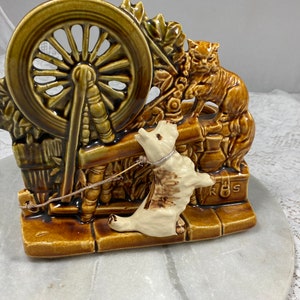 May include: A brown and green ceramic planter shaped like a spinning wheel with a cat and a dog figurine. The dog is white and brown and is pulling on a string attached to the spinning wheel. The cat is brown and is sitting on a box with the letters "R.S." on it.