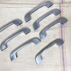 May include: Seven silver-toned cabinet door handles with a curved design. The handles are arranged in a staggered pattern on a white fabric background.