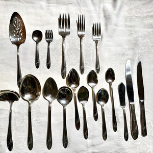 May include: A set of silverware, including forks, spoons, and knives, laid out on a white tablecloth. The silverware is made of polished silver and has a decorative design.