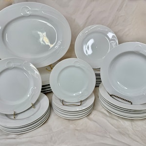 May include: A set of white porcelain dinnerware with a decorative swirl design. The set includes a large oval platter, a soup bowl, a dinner plate, and a salad plate.