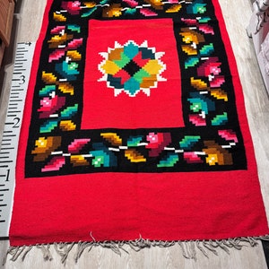May include: Red woven rug with a black border and a colorful floral design in the center. The rug has fringe on all sides.
