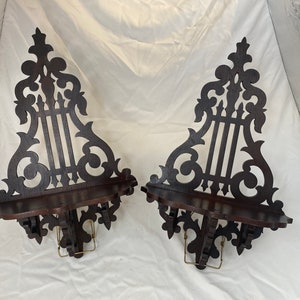 May include: Two ornate wooden wall shelves with intricate scrollwork and a harp-like design. The shelves are dark brown and have a slightly curved shape.