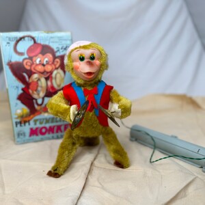 May include: A vintage green plush monkey toy with a red and blue vest plays cymbals. The monkey is wearing a white hat and has a wide smile. The toy is connected to a gray box with a red button. The box is labeled "Pepi Tuneful Monkey".