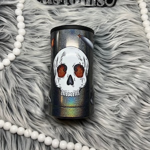 May include: A black and silver tumbler with a white skull design. The skull has red eyes and a black outline. The tumbler is on a white fluffy surface.