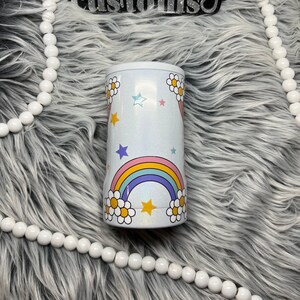 May include: White tumbler with a rainbow, stars, and daisies design. The tumbler has a glittery finish.