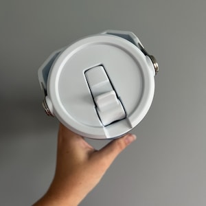 May include: A white and light blue insulated water bottle with a flip-top lid. The lid is white with a built-in handle and a silver metal clasp. The bottle is held by a person's hand against a gray background.
