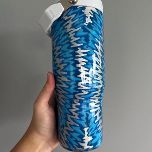 May include: A blue and silver insulated tumbler with a white lid and handle. The tumbler features a repeating pattern of blue and silver zigzags. The lid has a silver latch. The tumbler is held by a hand.