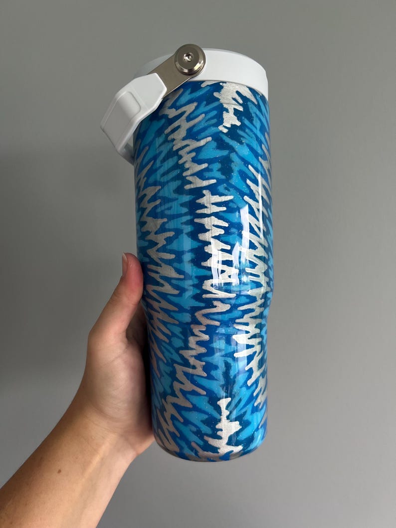 May include: A blue and silver insulated tumbler with a white lid and handle. The tumbler features a repeating pattern of blue and silver zigzags. The lid has a silver latch. The tumbler is held by a hand.