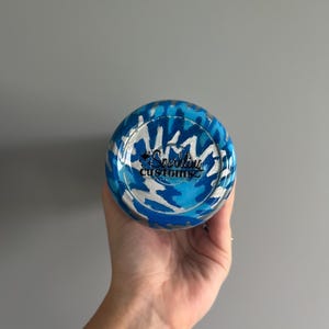 May include: A blue and silver tumbler with a camouflage pattern. The bottom of the tumbler features the text "Sparkling Customs" in a cursive font. The tumbler is held in a hand against a gray background.