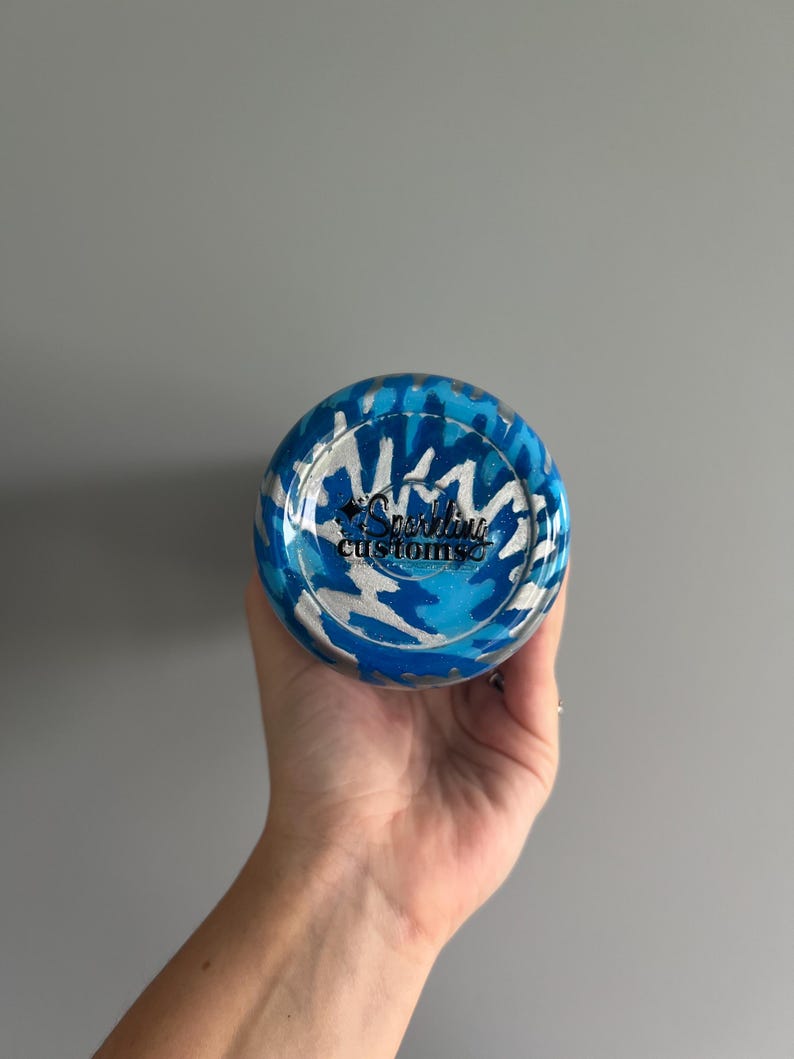 May include: A blue and silver tumbler with a camouflage pattern. The bottom of the tumbler features the text "Sparkling Customs" in a cursive font. The tumbler is held in a hand against a gray background.