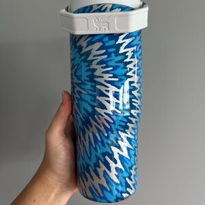 May include: A stainless steel tumbler with a white handle and lid. The tumbler features a blue and silver abstract pattern. The Stanley logo is visible on the handle. The tumbler is designed to keep drinks cold or hot.
