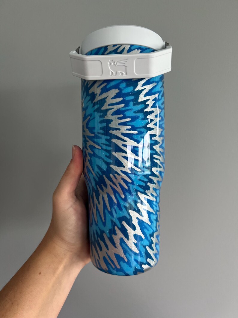 May include: A stainless steel tumbler with a white handle and lid. The tumbler features a blue and silver abstract pattern. The Stanley logo is visible on the handle. The tumbler is designed to keep drinks cold or hot.