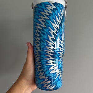 May include: A blue and silver insulated tumbler with a white lid. The tumbler features a repeating pattern of silver lightning bolts against a blue background. It has a handle and a white top.