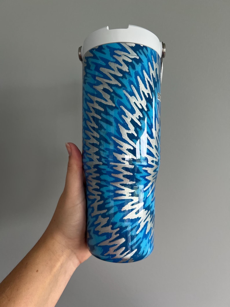 May include: A blue and silver insulated tumbler with a white lid. The tumbler features a repeating pattern of silver lightning bolts against a blue background. It has a handle and a white top.
