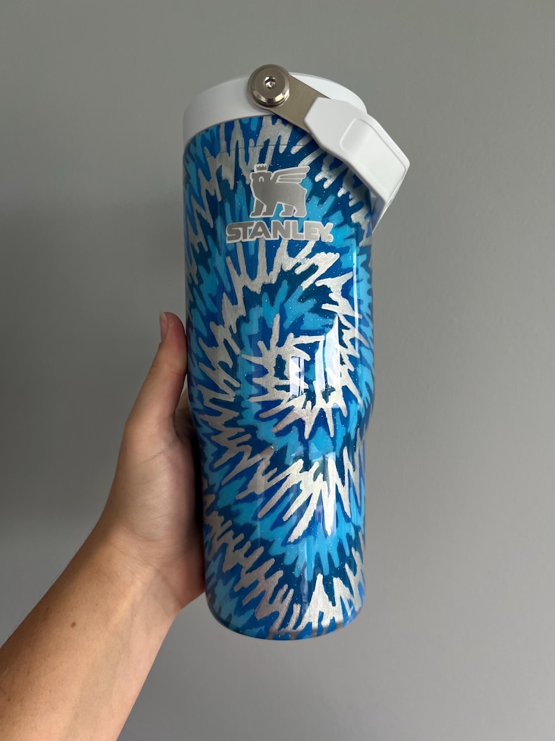 May include: A blue and silver Stanley tumbler with a white handle and lid. The tumbler features a tie-dye pattern and the Stanley logo. The tumbler is designed to keep drinks cold or hot.