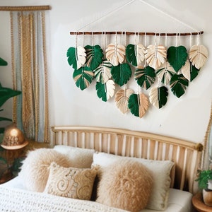 Op de afbeelding: Een macramé wandhanger met groene en crèmekleurige bladeren, hangend boven een bed met witte beddengoed en een houten hoofdeinde. De wandhanger is gemaakt van garen en heeft een houten lat aan de bovenkant.