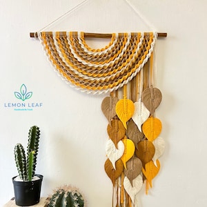 May include: A macrame wall hanging with a half-circle design in shades of yellow, brown, and white. The hanging features a cascade of woven leaves in the same colors.
