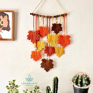 Pode incluir: Uma tapeçaria de parede feita de macramé com um ramo castanho e várias folhas laranja, amarelas e castanhas. As folhas são feitas de fio e têm uma franja na parte inferior. A tapeçaria de parede está pendurada numa parede branca com duas plantas em vasos em primeiro plano.