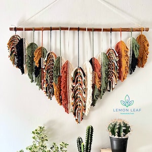 以下が含まれることがあります： 木製の棒の上部にマクラメで作られた壁掛け。マクラメは羽の形をしており、黒、白、緑、オレンジ、茶色など様々な色で作られています。壁掛けは白い壁に掛けられており、その前に2つの鉢植えがあります。