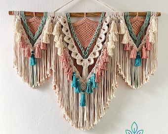 May include: A macrame wall hanging with a geometric design in shades of peach, teal, and cream. The hanging is made of cotton cord and has a wooden dowel at the top.