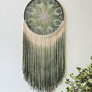 Puede incluir: Un atrapasueños de color verde y crema degradado con un diseño de ganchillo y flecos. El atrapasueños está colgado de un cordón negro contra una pared blanca.