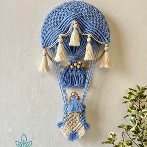 Peut inclure: Une suspension murale en macramé en forme de montgolfière bleue et blanche avec une petite poupée suspendue en bas. La montgolfière a un anneau en bois et des perles en bois.